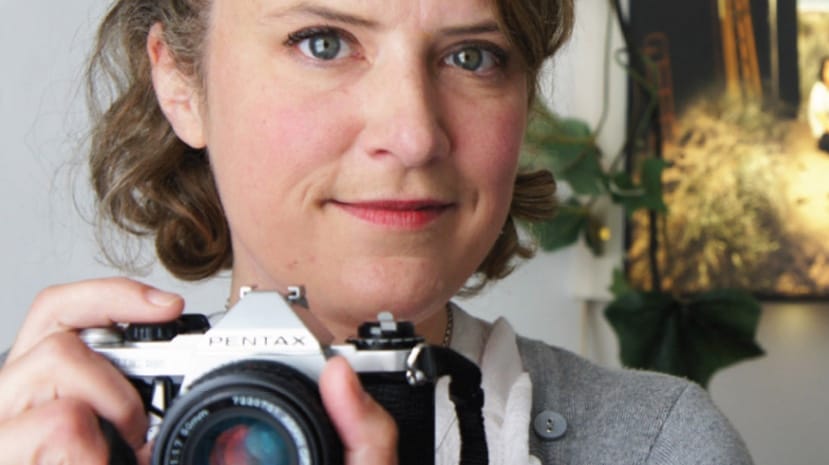 A white woman with green eyes and medium length curly hair faces the camera with a smile. She is pointing her own camera at the camera taking the picture.