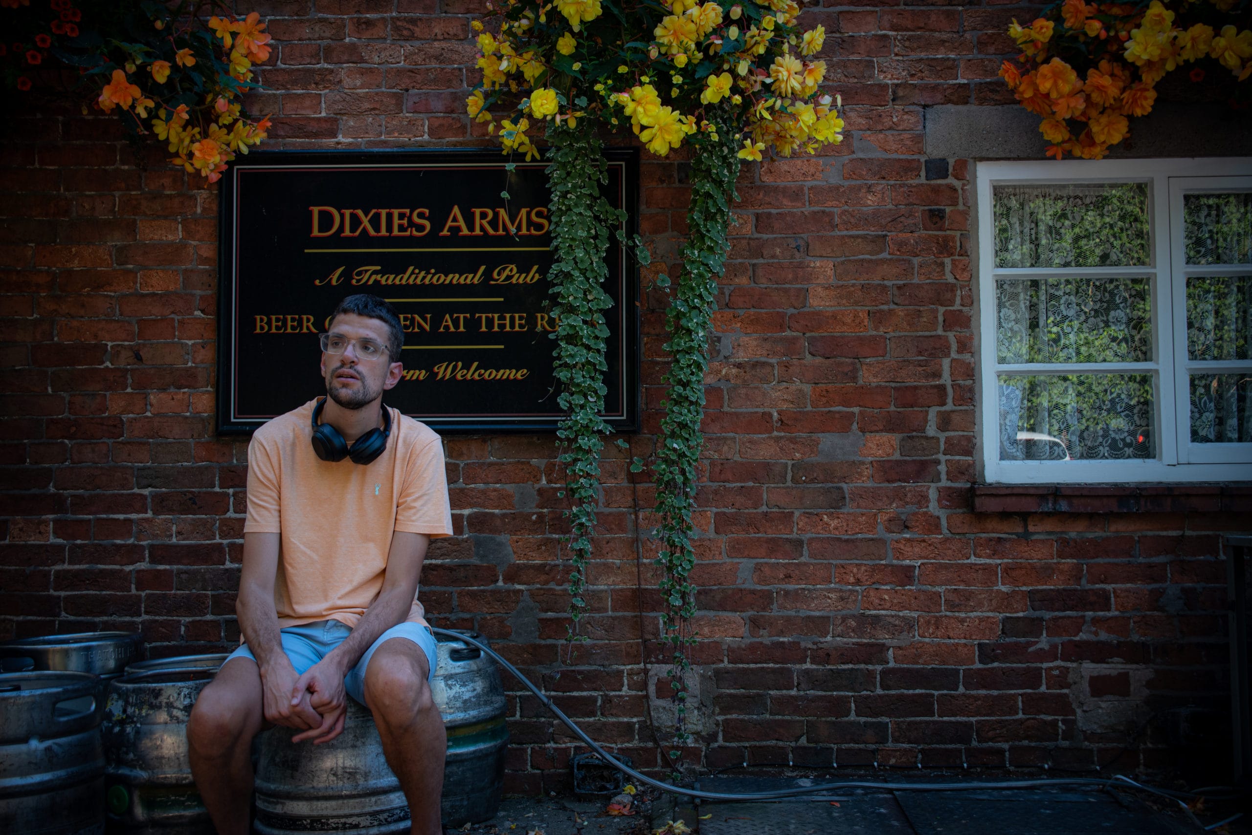 Neal is siting on a beer keg outside a pub called 'Dixies Arms'. He is wearing glasses, a yellow t-shirt, blue shorts and has headphones around his neck. There are hanging baskets full of yellow flowers above his head.