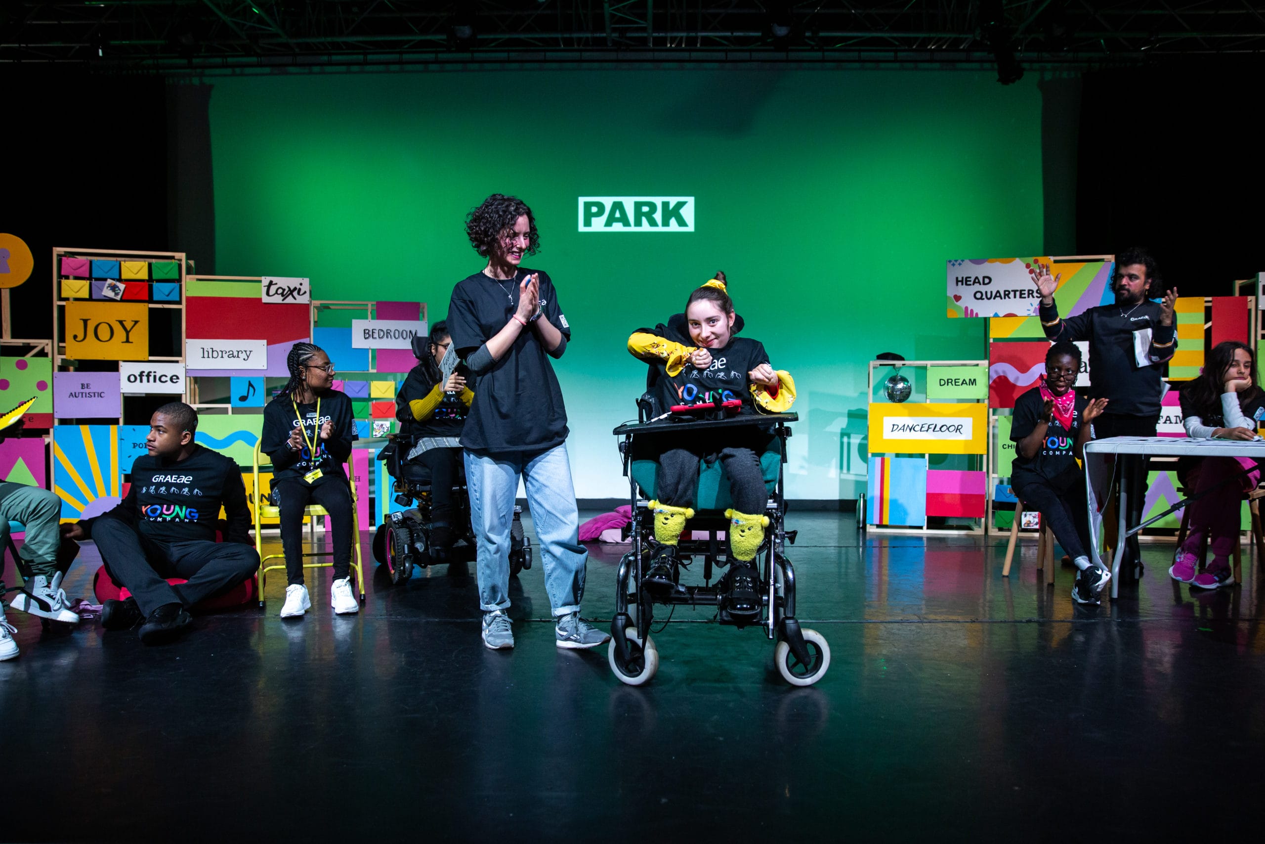 A performer in a wheelchair is centre stage, surrounded by other performers who are clapping along.