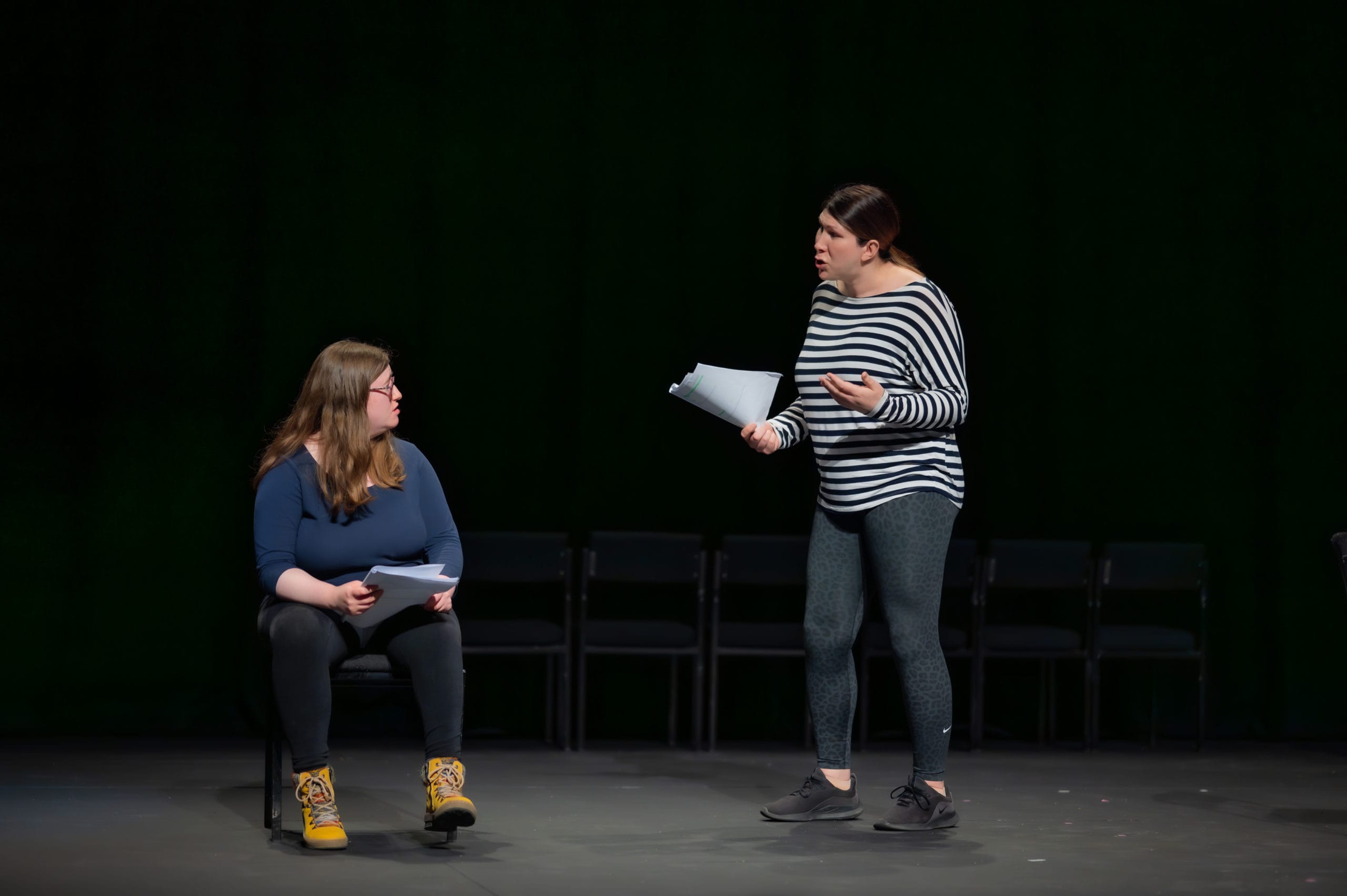 Two performers on stage, holding scripts. One of them is sitting down, the other is standing, looking towards their stage partner and speaking.