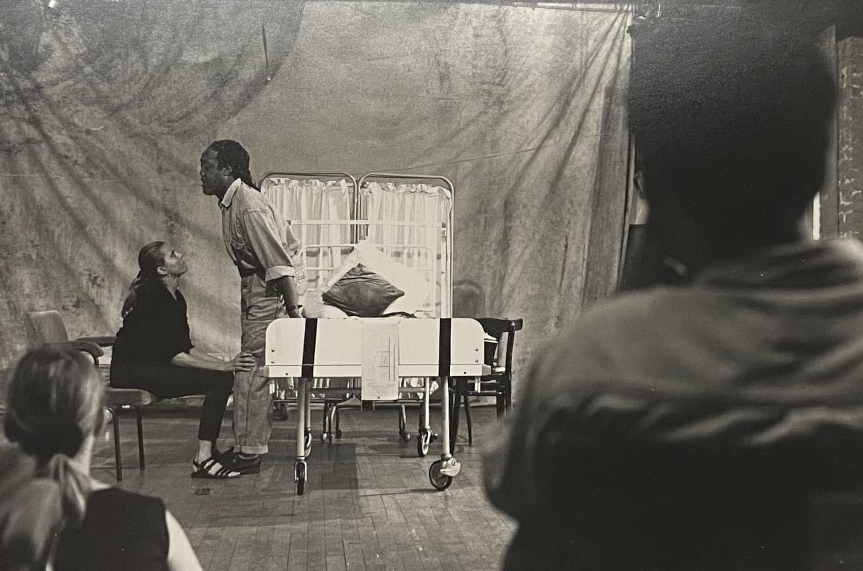 Two performers on a stage set as a hospital. One sits in a chair, holding the knees of the other, who is standing over them.