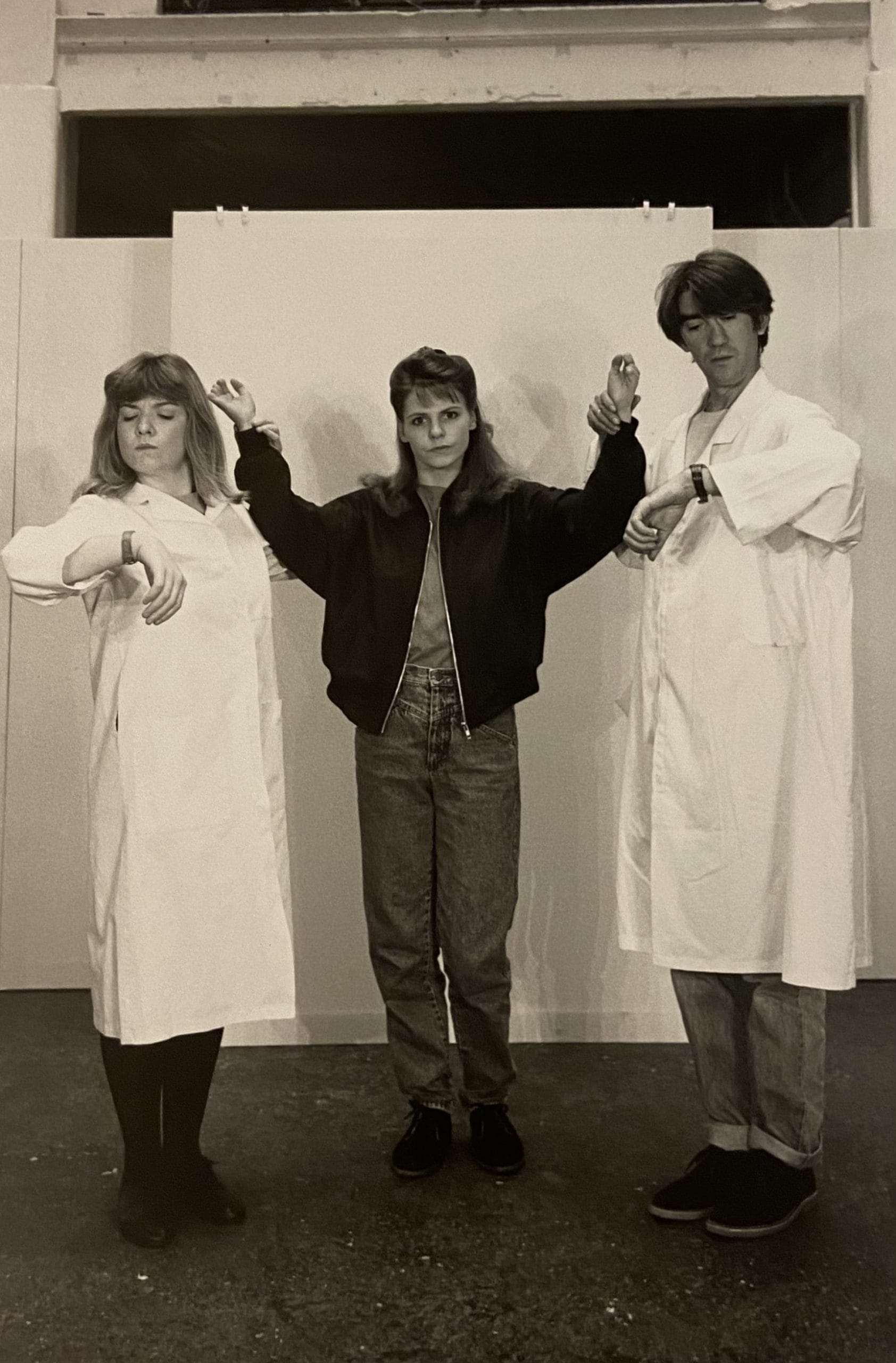 Three performers on stage, two wearing lab coats and looking at their watches. The third stands in the middle with her hands up in the air.