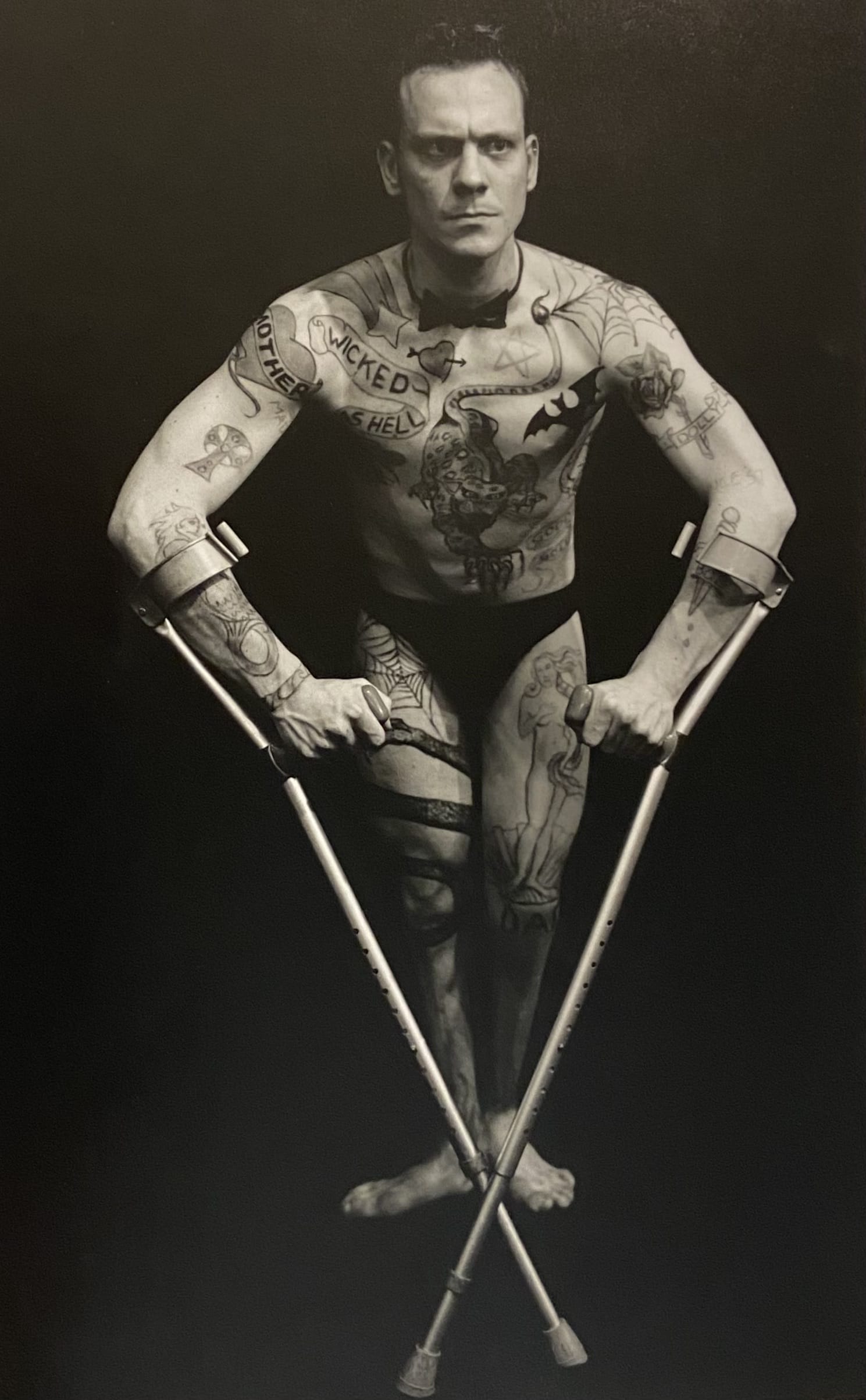 A male performer, covered in tattoos and wearing black underwear, flexes whilst leaning forward onto his crutches.