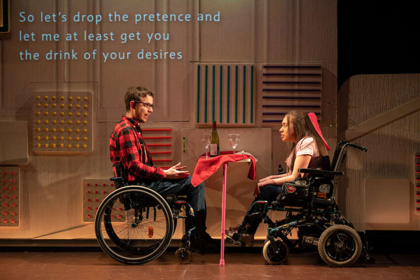 Two performers on stage, sit across from each other at a restaurant table. They are both using wheelchairs, hers in motorised, his is not. The man is speaking and his lines are projected onto the wall behind him. He is saying 