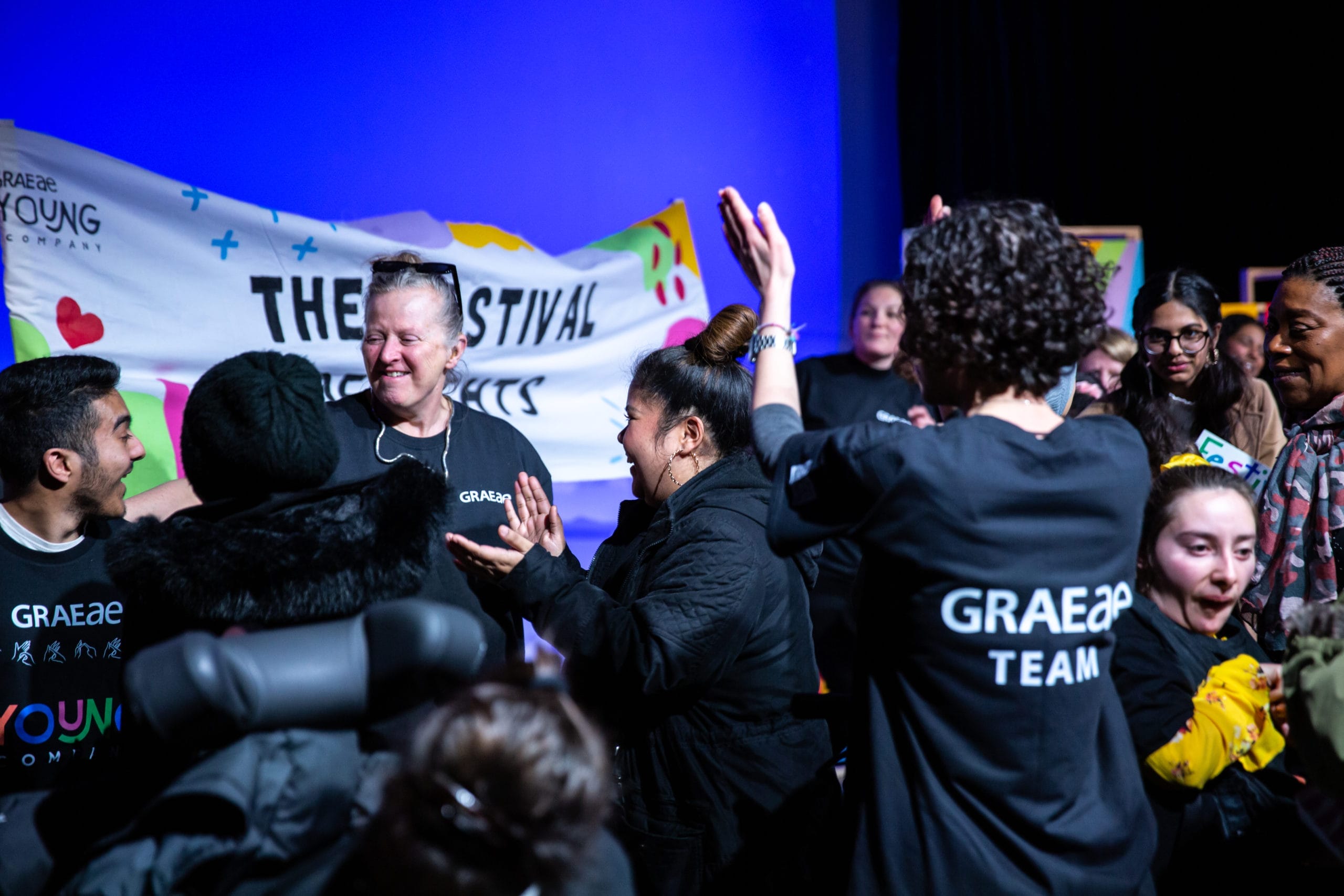 A crowd of people and performers all wearing t-shirts reading 'Graeae Team'. They are clapping and cheering.