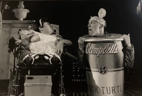 Two performers on stage, One of them is sitting in a wheelchair, the other is dressed as a giant campbells soup can.