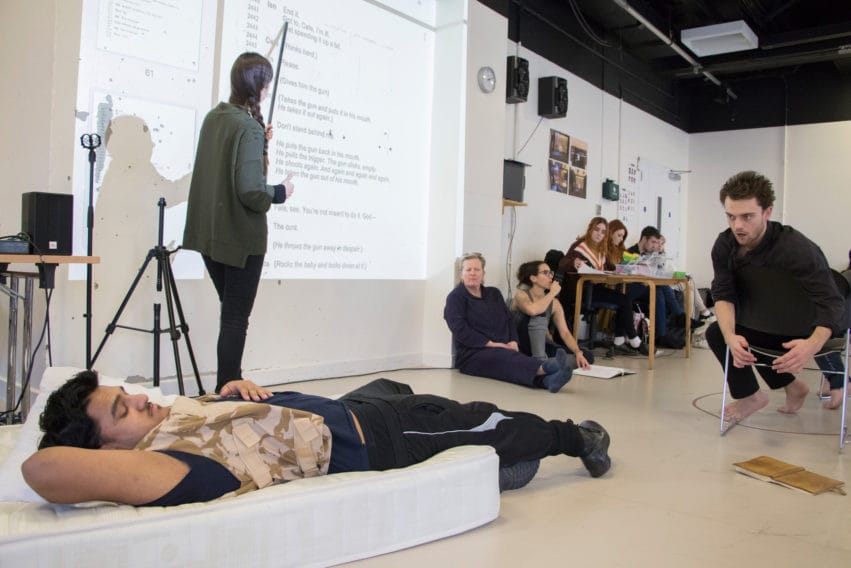 A rehearsal, on man lays on a mattress whilst another cranes over to talk to him. People observe in the background.