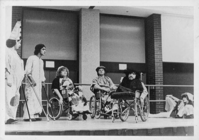 Performers on stage. Three are in wheelchairs, one is lying on the floor, and two a stood up. All appear to be looking at the same fixed point in the distance.