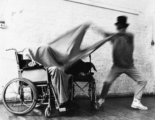 A black and white blurry action shot; someone in a top hat quickly pulls a large blanket off of someone in a wheelchair.
