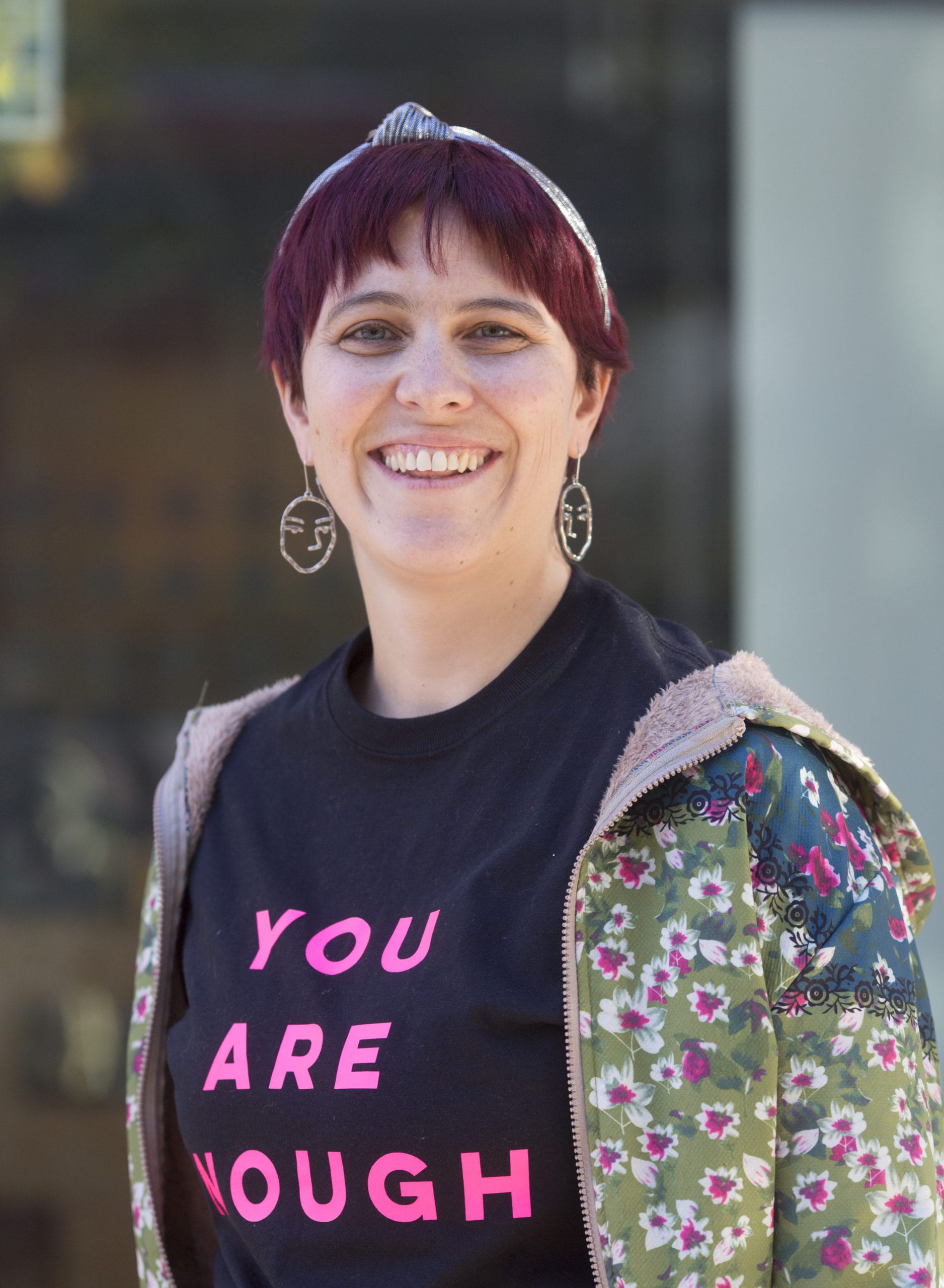 Image of a white woman with redish-purple hair smiling at the camera. She is wearing a headband and earrings. She has on a black shirt that says, "YOU ARE ENOUGH".