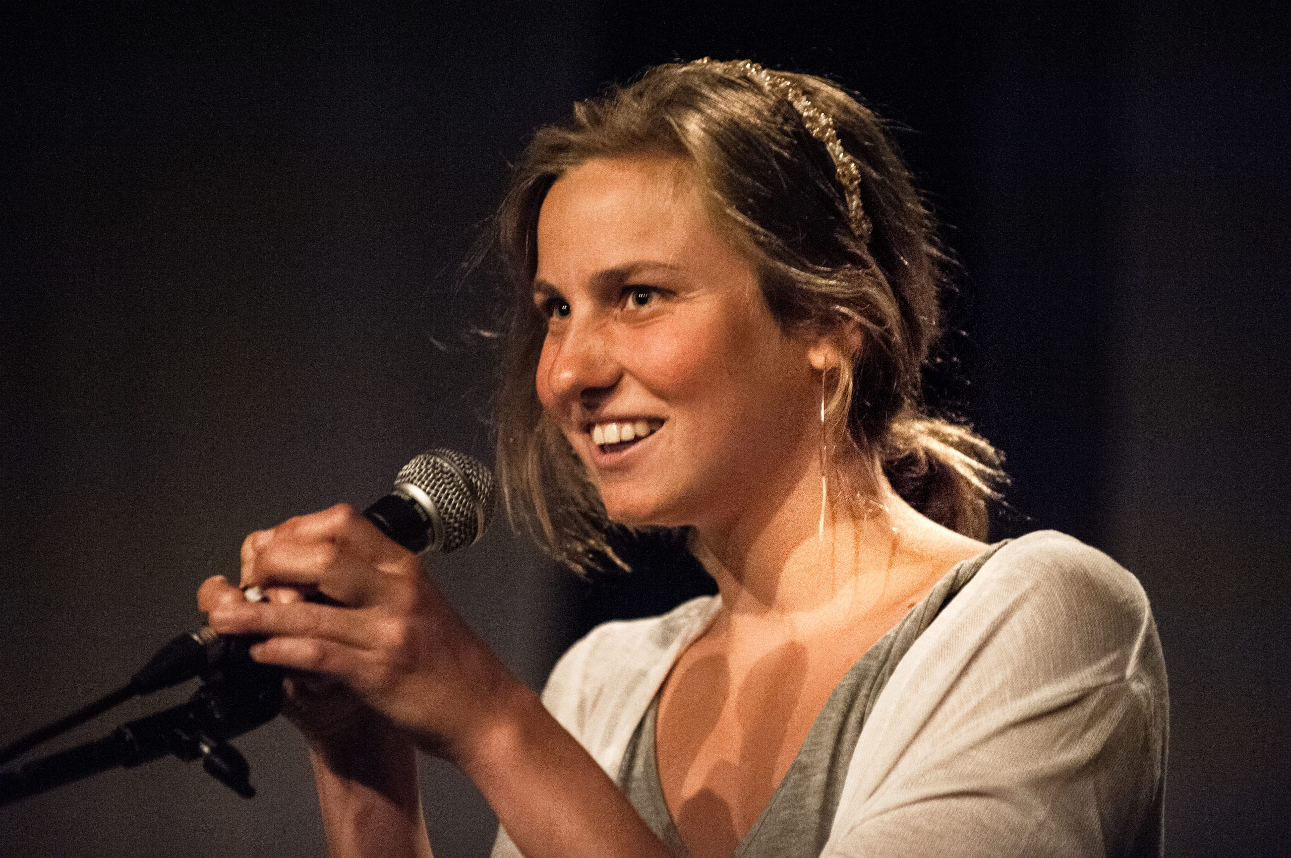 Colour photo of an actor speaking into a microphone. Their hair is pulled back, they are wearing a white cardigan, and a grey dress.