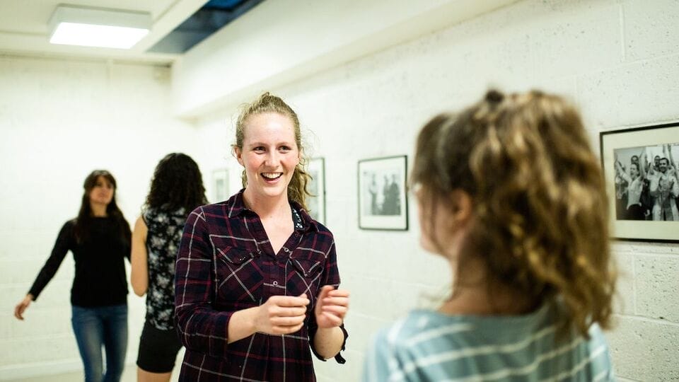 A young woman stands in conversation with another woman with her back to the camera. Gallery images appear on the wall behind.