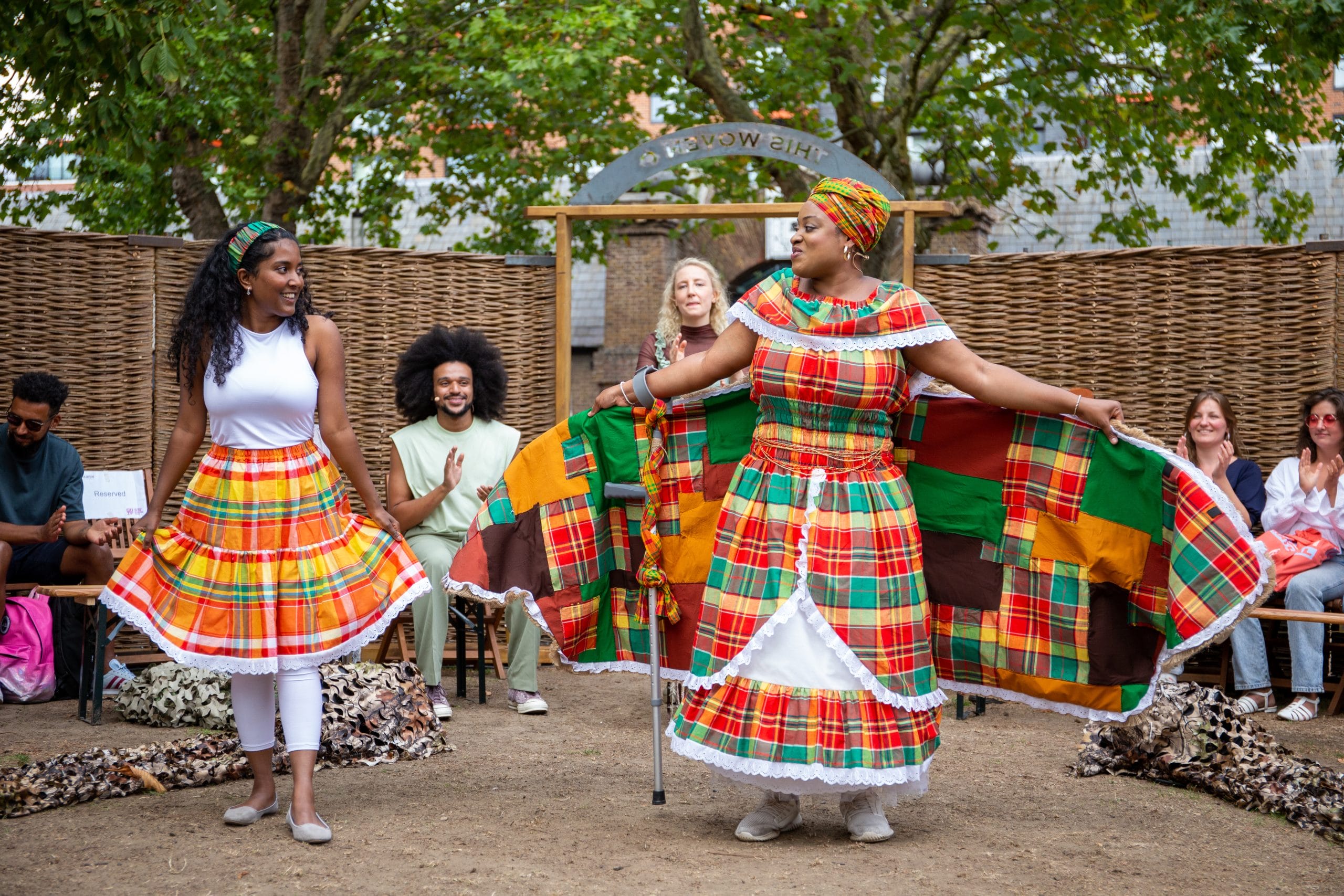 Two women in colourful costumes are standing in the middle of a semi-circle of people.