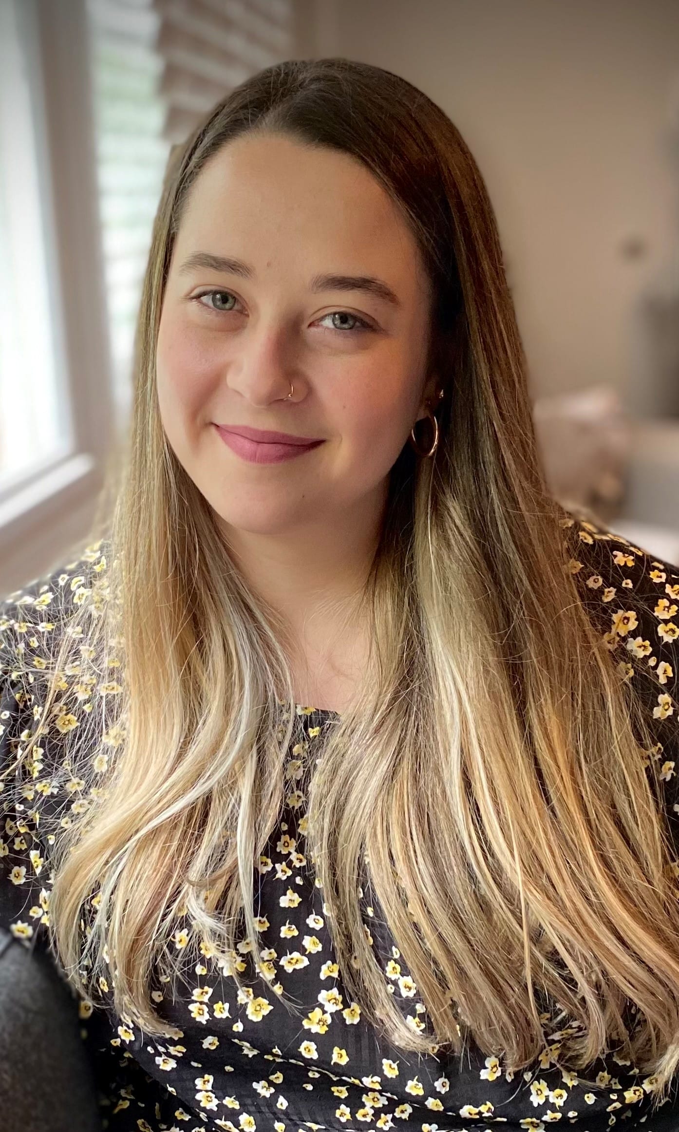Image of woman with long brown-blonde hair. She is wearing a floral top and she is smiling;
