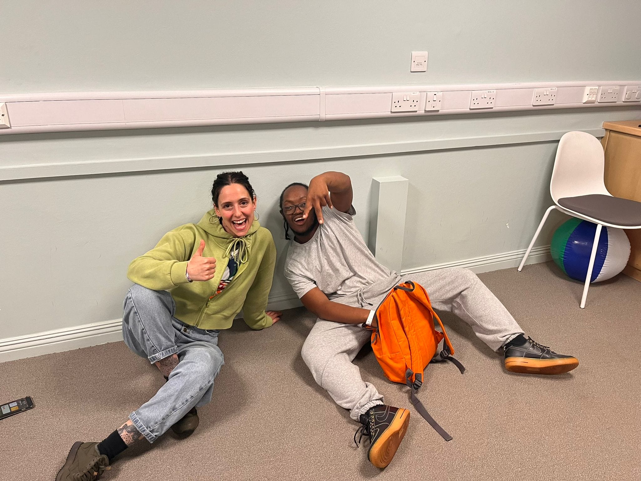 Stella and Marcus sitting together on the floor. Stella is wearing a green hoodie and jeans. Marcus is wearing a grey shirt, grey joggers, and is holding an orange backpack.