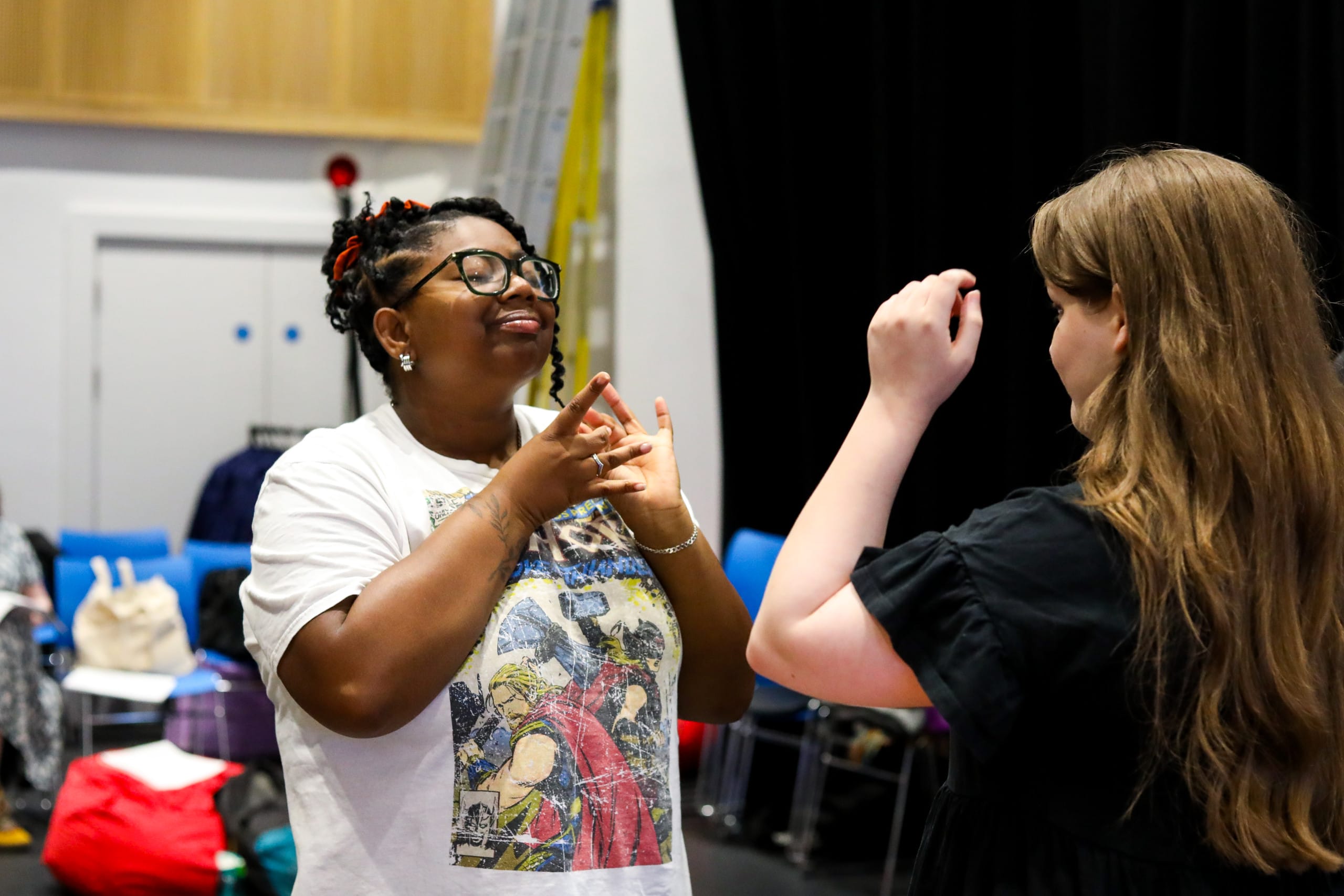Photo of Cherie, she is a black woman, she is wearing glasses and a white graphic t-shirt. She is standing across from another performer and is signing.