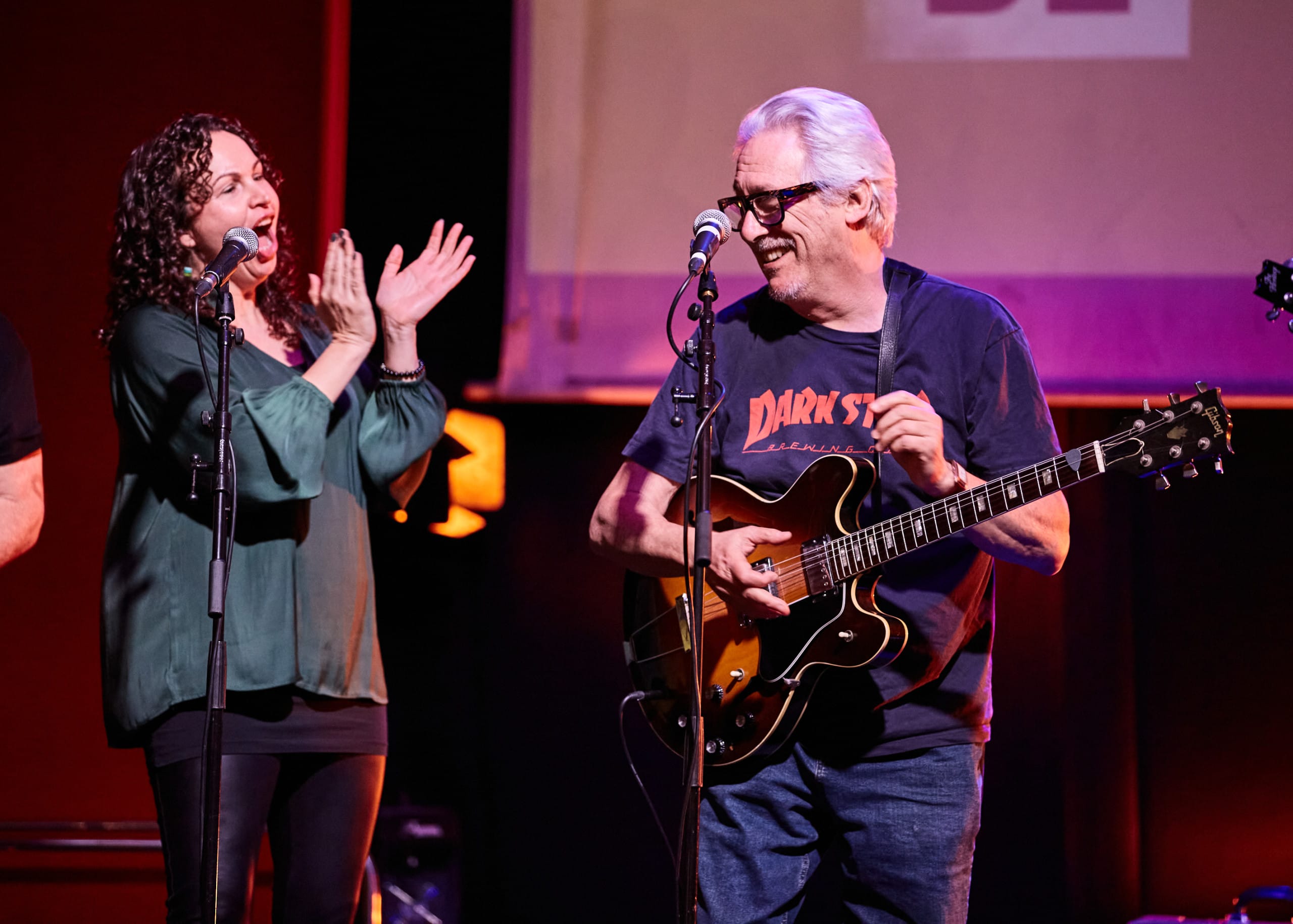 Jude and Chaz are performing a song. Jude is a white woman in a green blouse and dark trousers. Chaz is a white man, wearing a t-shirt and jeans, he is playing the guitar.