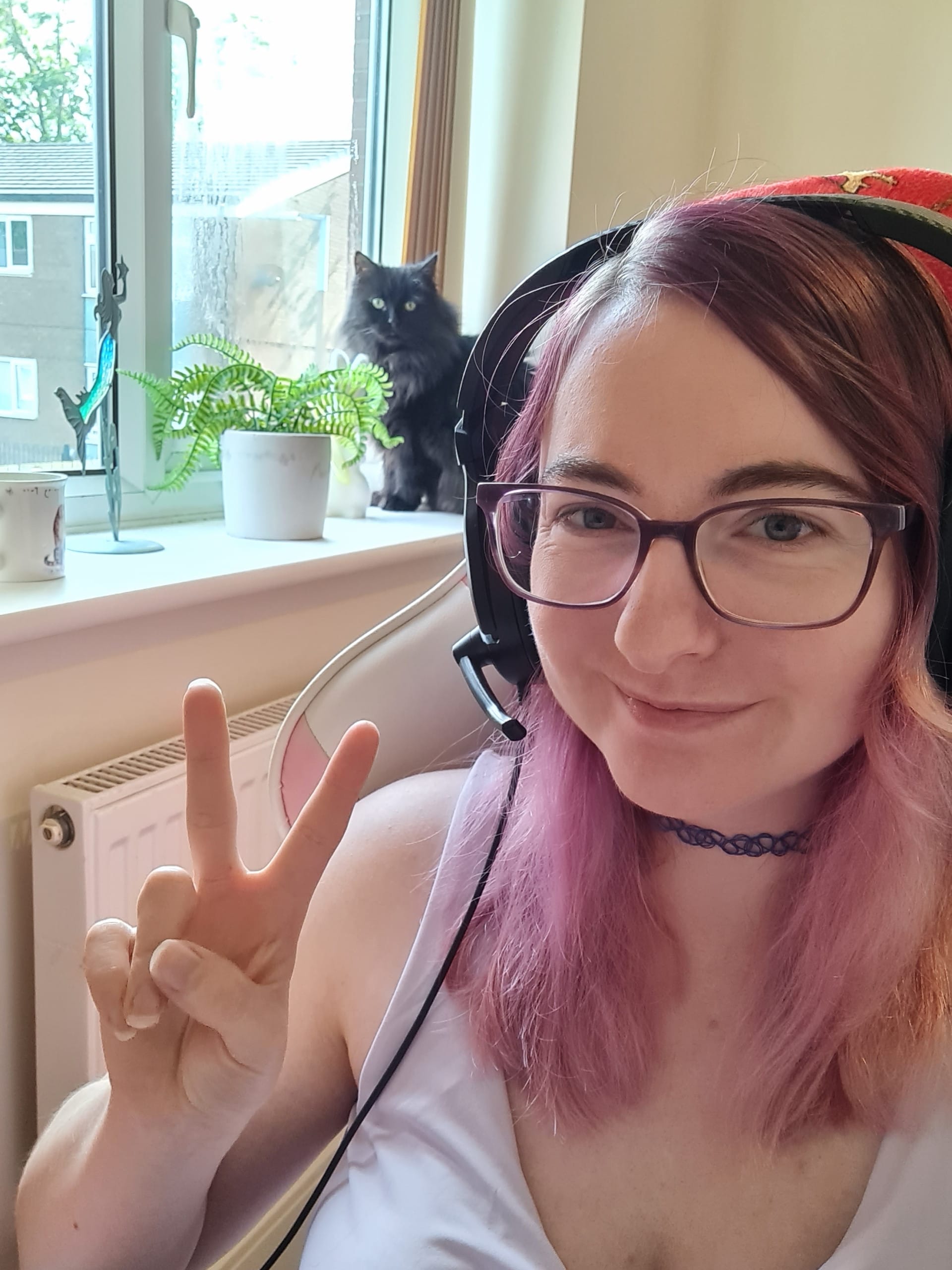 'A white woman making a peace sign. She is wearing glasses and a gaming headset. She has dyed purple hair. In the background is a fluffy black cat