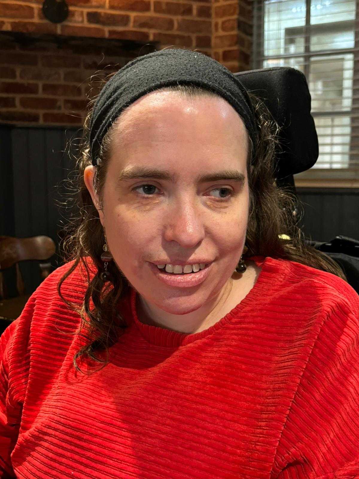 A white woman in a red jumper. She has dark, shoulder length hair and is wearing a black headband.