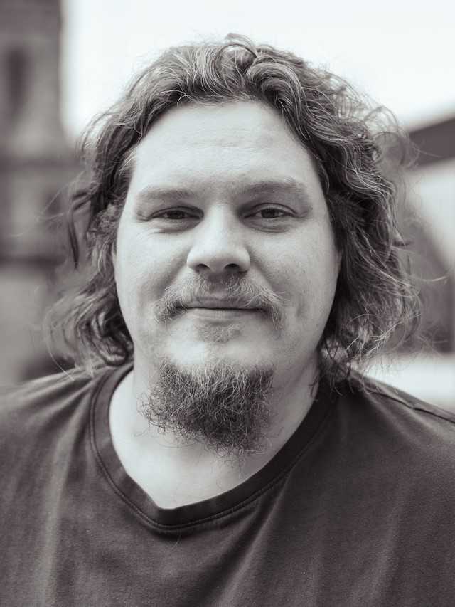 Black and white image. A male in his thirties. He has a rounded face and full checks. He has shoulder length curly dark brown hair, a goatee beard, and a moustache.