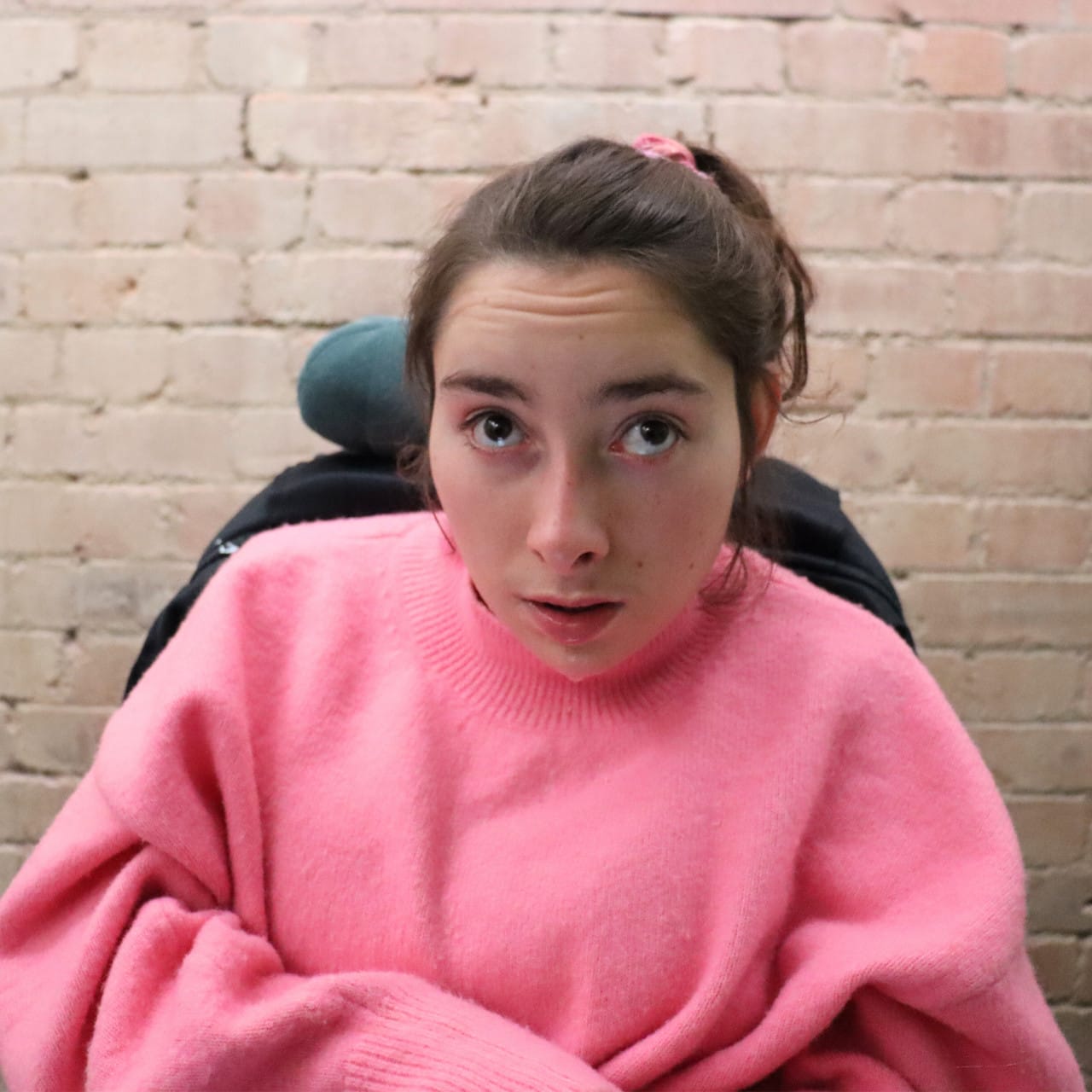 A young white woman with brown hair and brown eyes, wearing a pink jumper.