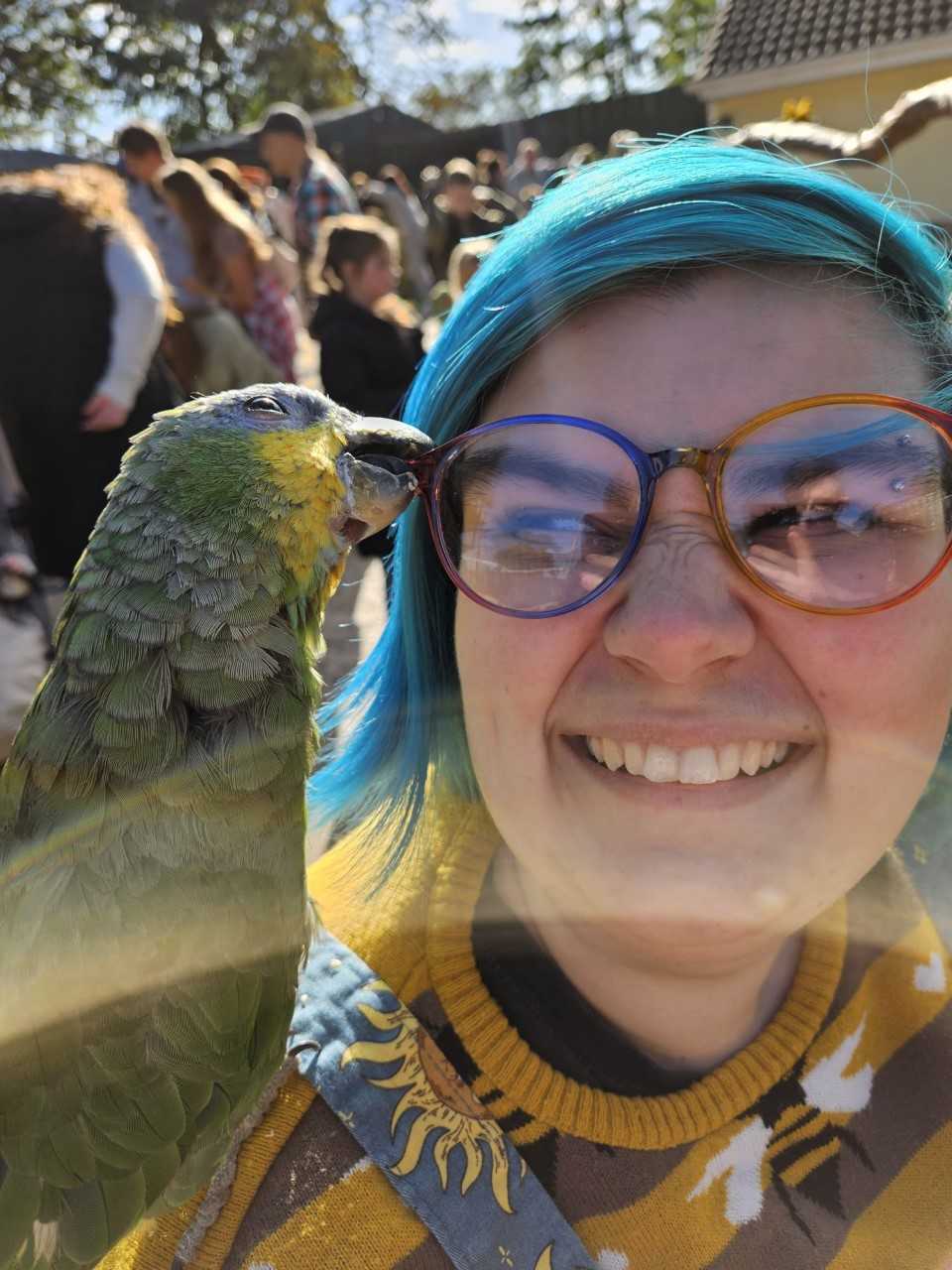A photograph of Katie, a fat, white, blue haired person, smiling in the sun. A parrot sits on their left shoulder, attempting to steal their glasses, which are round with rainbow frames.