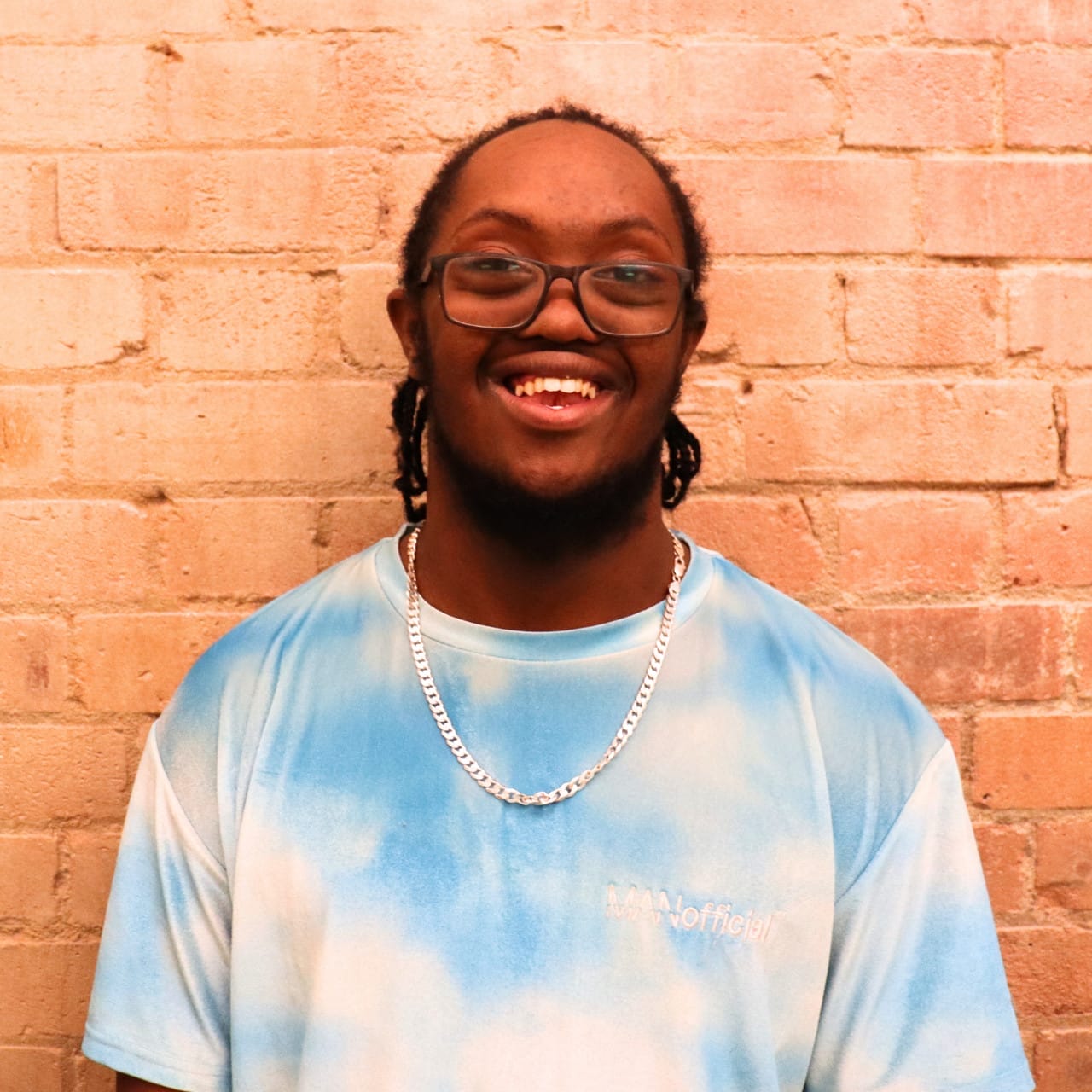 A black young man with a beard, wearing glasses, with big hair in twists, wearing a metal chain, and blue t-shirt.
