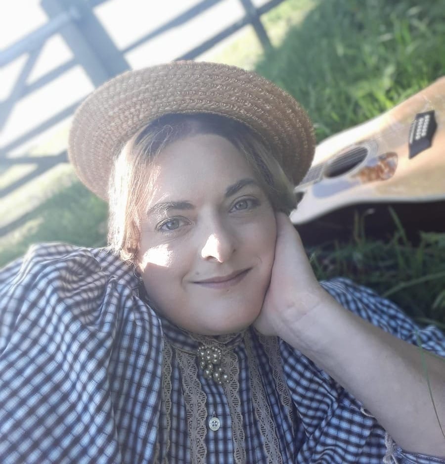 Pixie, a white person in their early 40s, lays with a guitar in a grassy field in strong sunlight. They are wearing a straw hat and checked blouse with a pearl brooch
