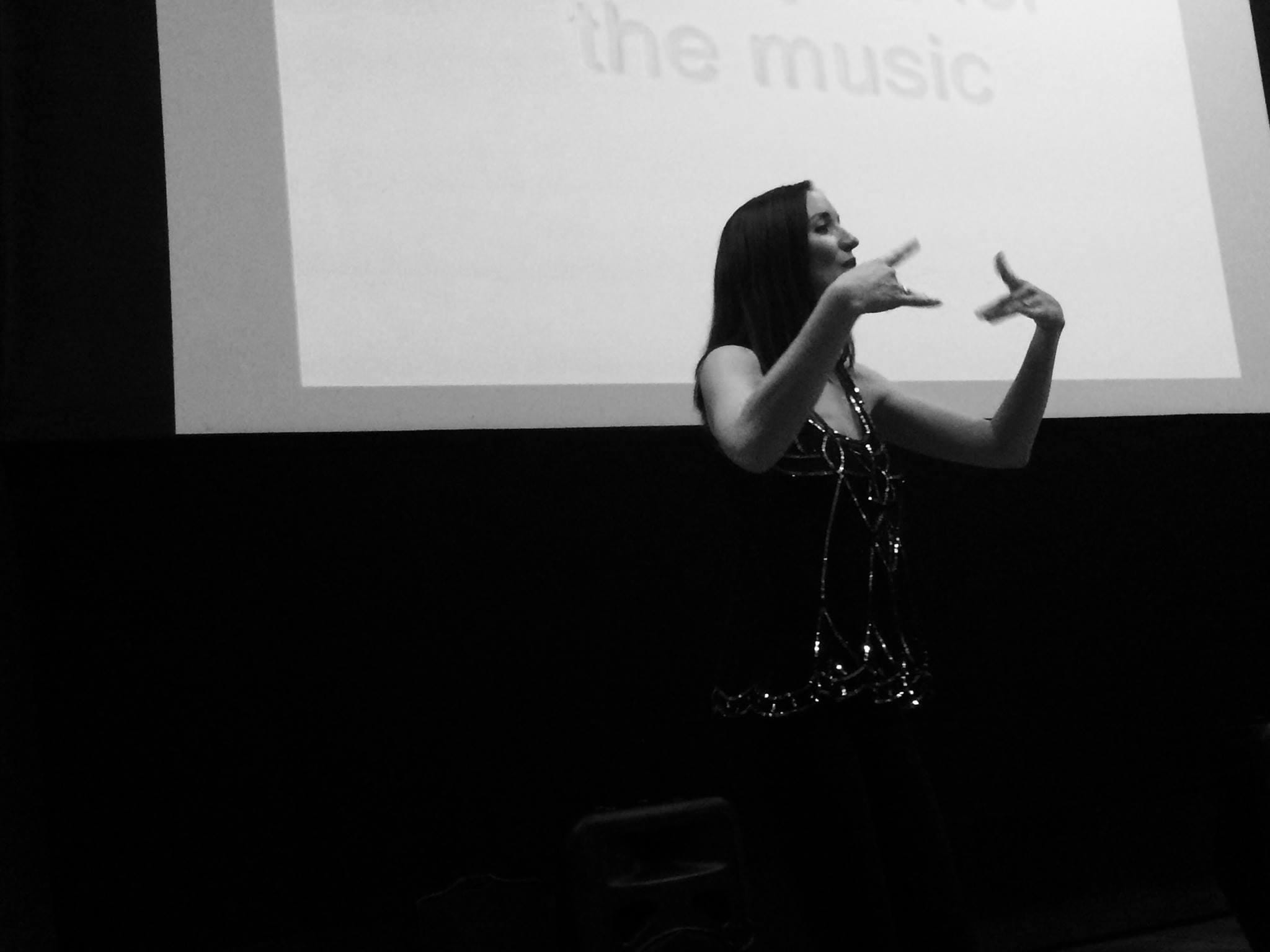 A black and white image of a white female with long dark hair and a sparkly top, signing to an audience with both hands in front of her