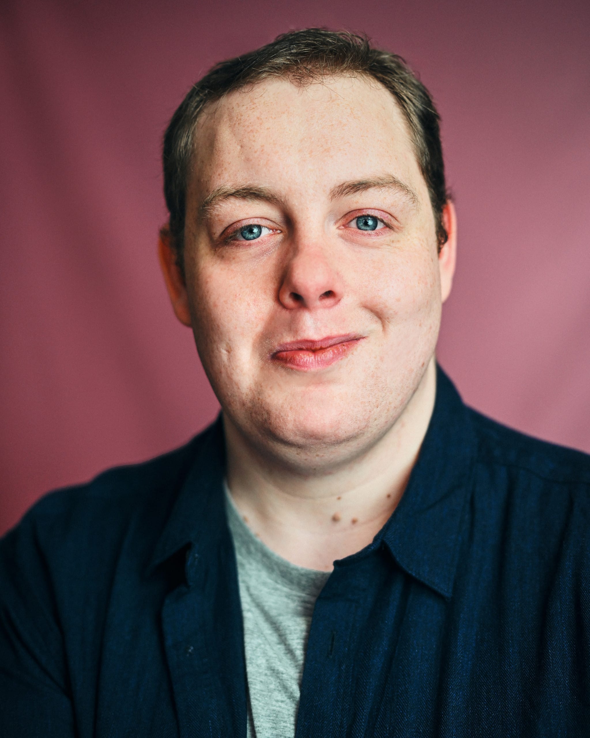 Young white man; with brown hair; blue eyes and stubble amongst his many freckles, he boasts a cheerful grin and is wearing a dark blue shirt over a green t-shirt.