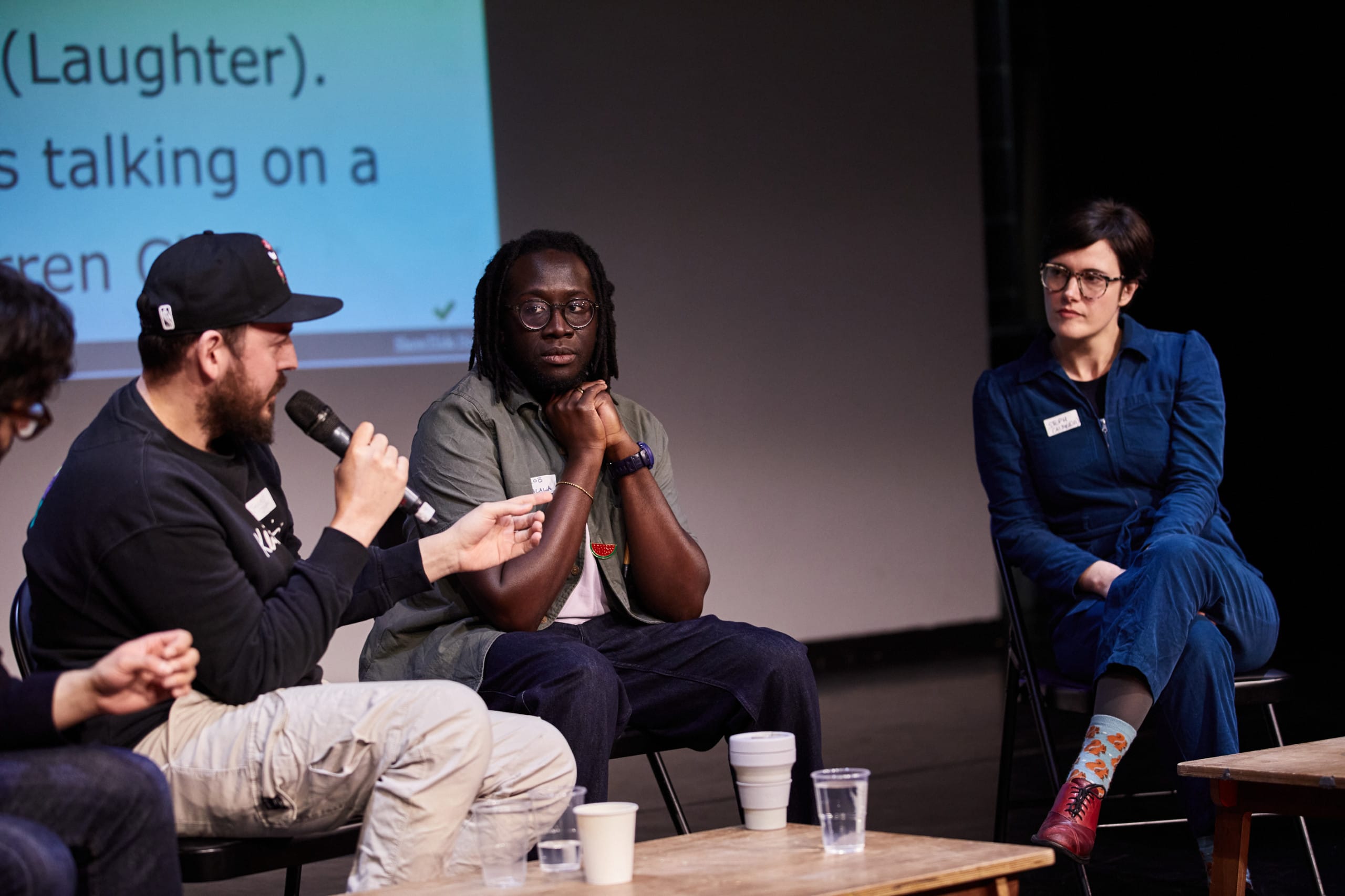 Panel presenation, three individuals in shot. A white man, wearing a baseball cap and a jumper is holding a microphone, speaking. The others are looking at him.