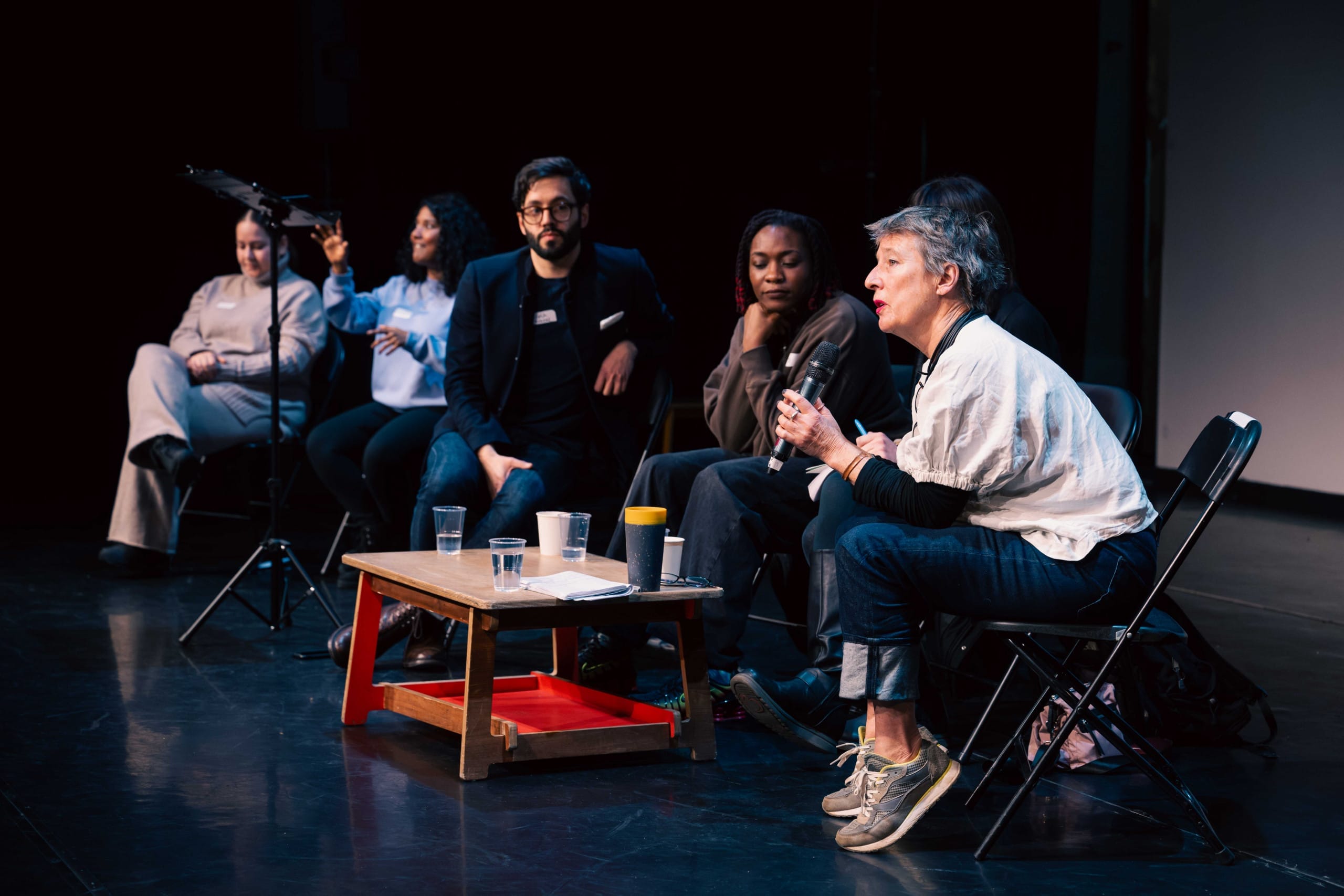 Image of a panel discussion. Six individuals are sat on a stage, on the end there is a woman with short hair holding a microphone, she is speaking while the others look at her.