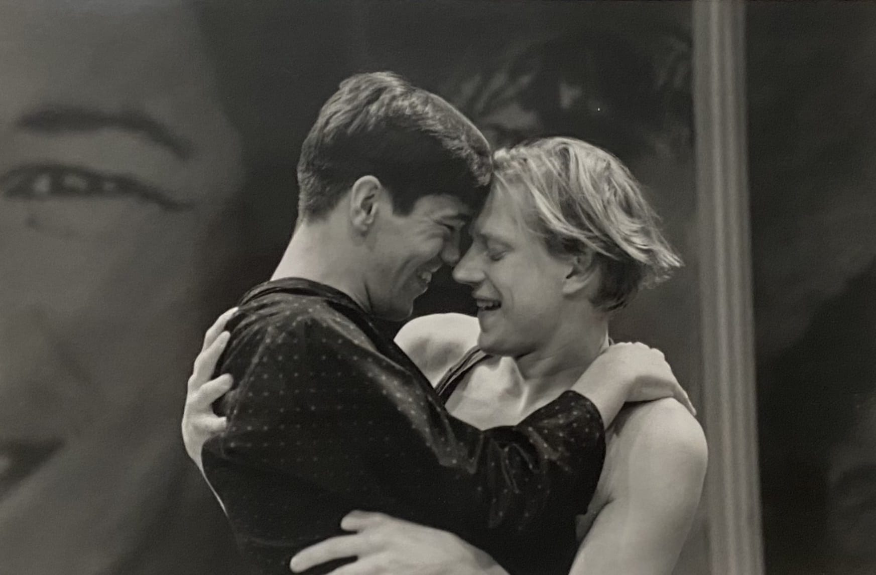 Black and white production photo from a kind of immigrant. Two men are embracing one another and smiling.