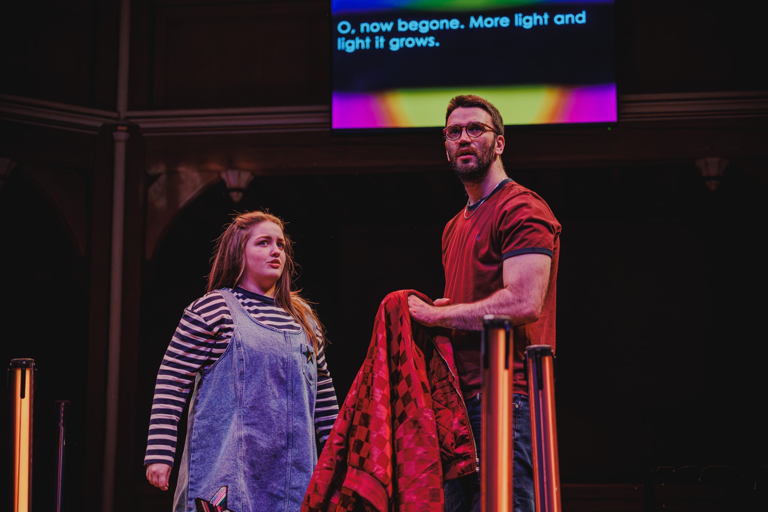 Ciaran Forrest in a red tshirt holding a red jacket looks out into the audience, shocked. He stands next to another actor wearing blue dungarees