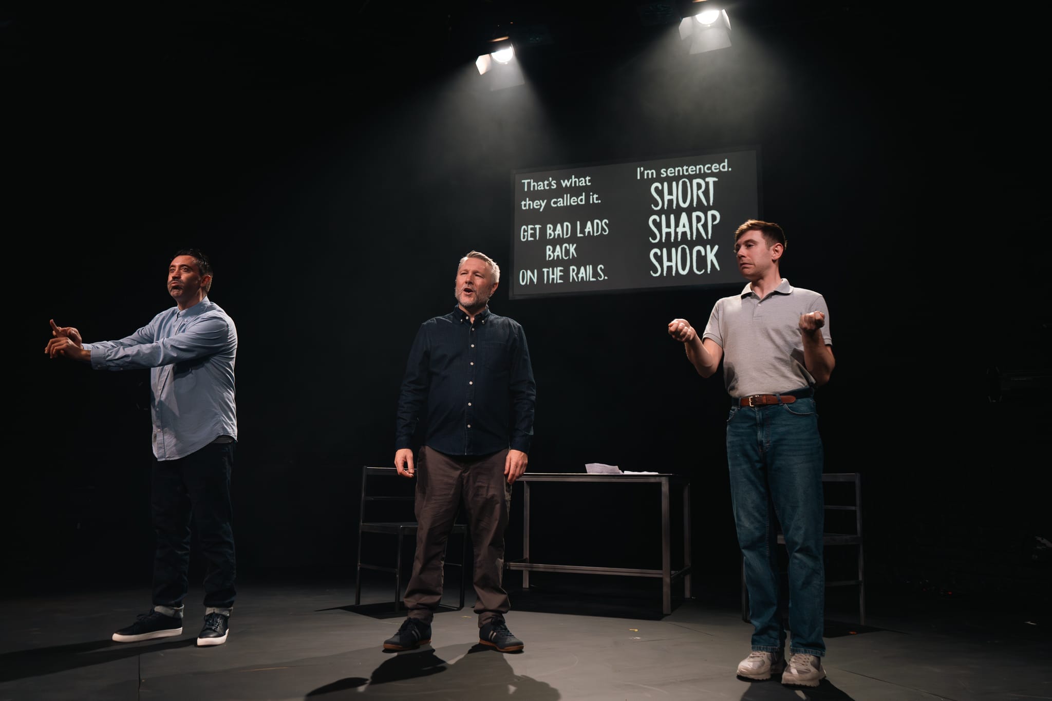 Three cast members stand on stage, one signs . The caption screen behind reads short sharp shock
