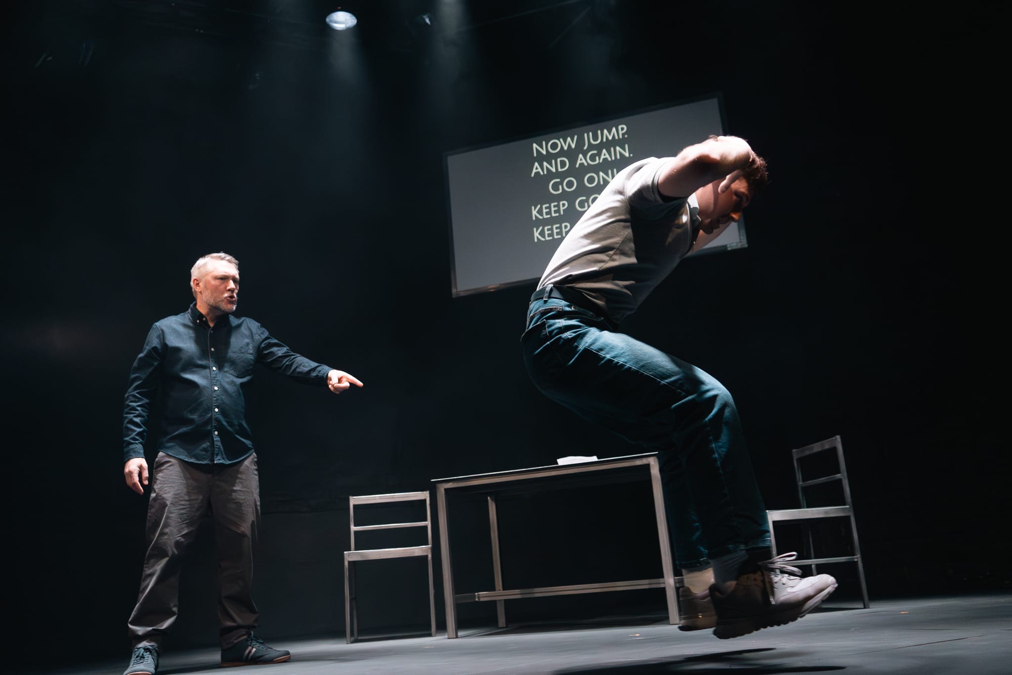 Cast member Danny Raynor stands and points shouting at cast member Robin Paley Yorke as he jumps around the stage.