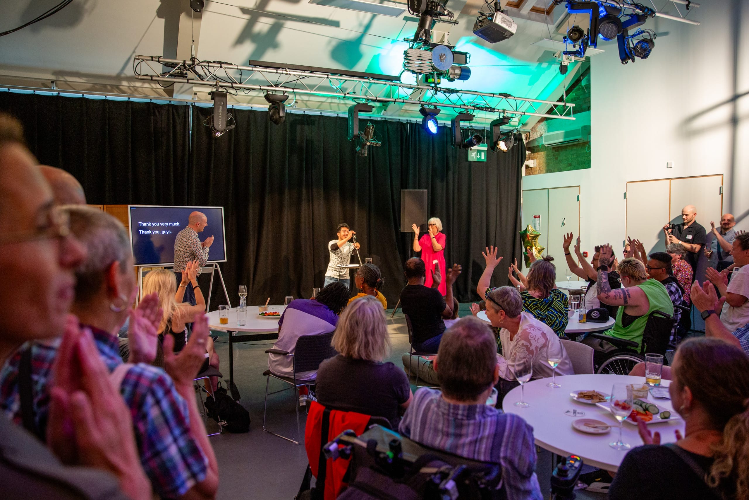 A room full of people in Graeae's rehearsal room watching and listening to a young person talking