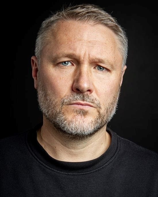 A white man with a grey beard and grey hair wearing a black t shirt.