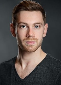 A white man with short hair and a moustache and beard wearing a grey t shirt