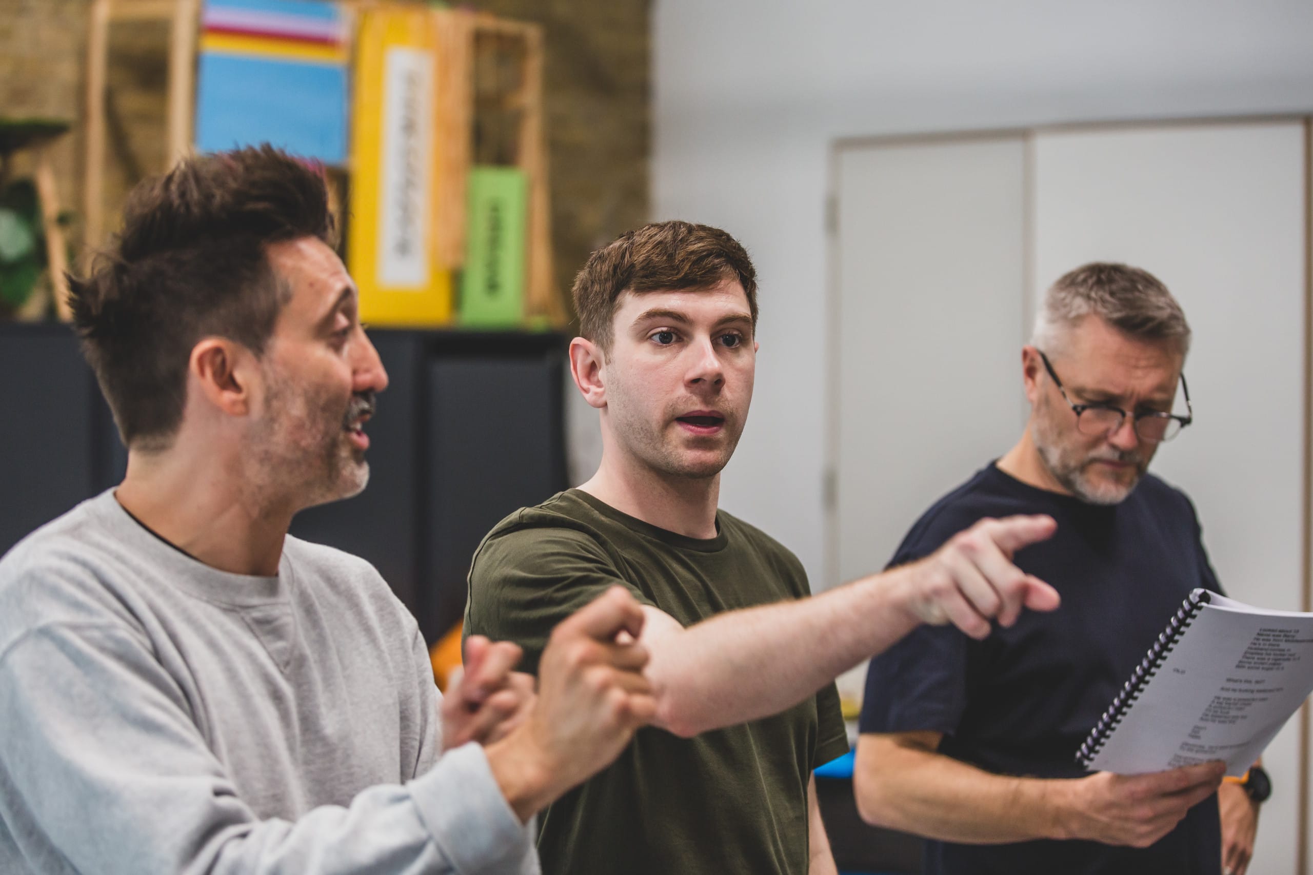 Cast members all stand in a line, Craig signs, Robin points his finger and Danny is reading his script.