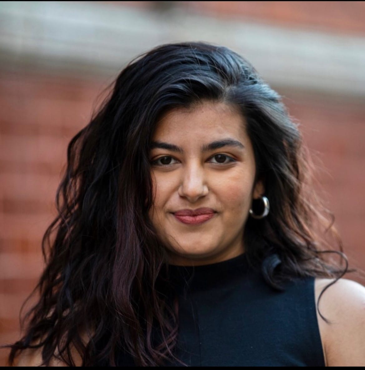 Brown woman with shoulder length black hair, brown eyes, light red lipstick, smiling and wearing a black top with silver earrings.
