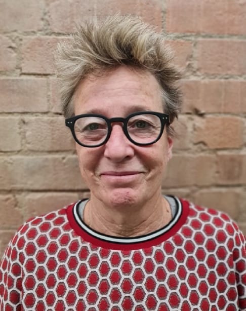 Hetty, a white woman with short blonde hair, wearing black glasses a red and white jumper against a brick wall