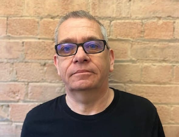 Laurence, a white man with short grey hair wearing glasses with a black top against a brick wall
