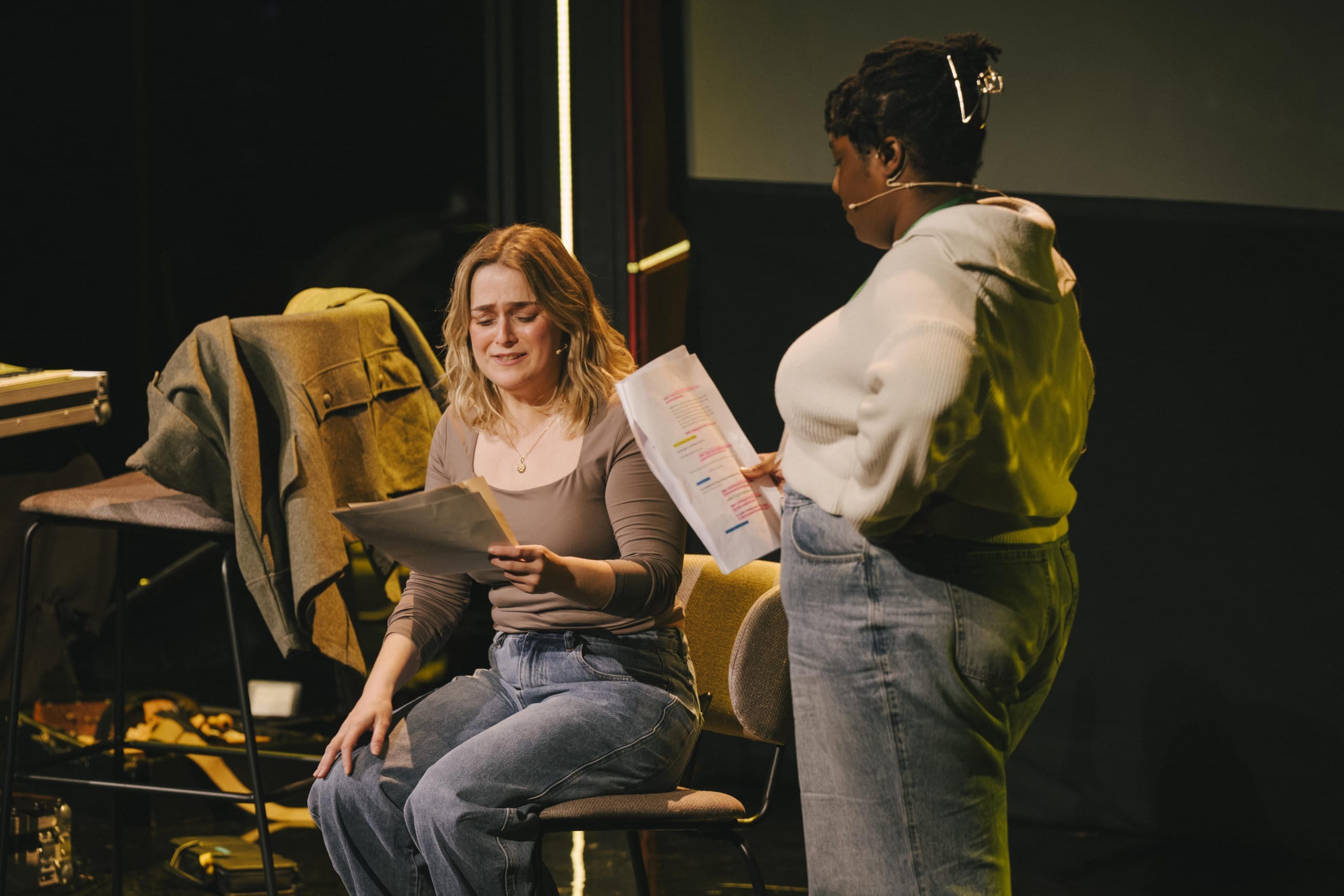 One person sits and one stands beside holding scripts on stage.